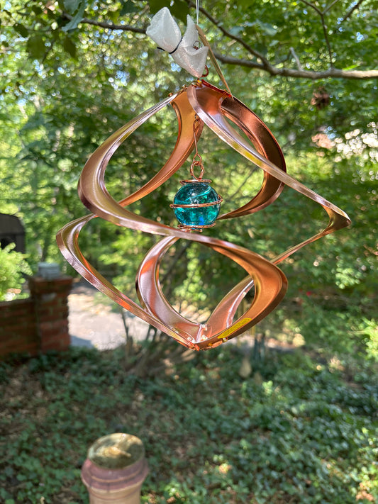 copper-colored metal wind spinner with a central blue-green glass ball, hanging outdoors in front of a blurred green garden background